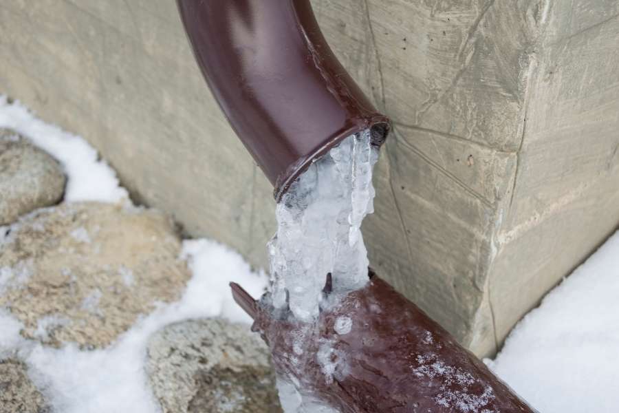 A close-up of a frozen brown metal downspout overflowing with thick icicles during winter.